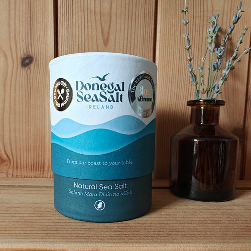 A cylindrical container of Donegal Sea Salt Ireland natural sea salt sits on a wooden surface against a wooden plank wall, styled next to a small dark amber glass vase with dried lavender sprigs.
