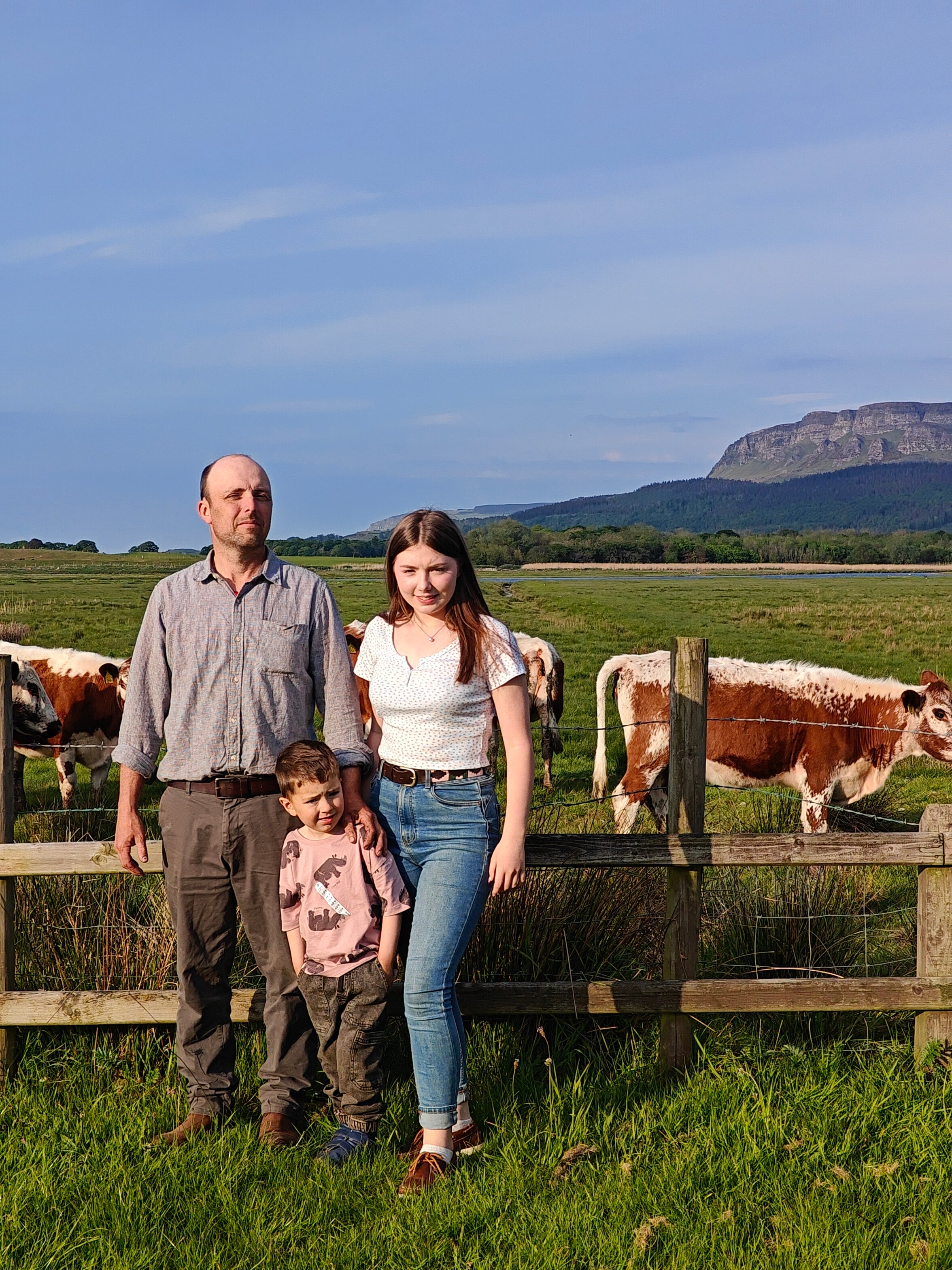 Family of three standing in a field with cows and mountains in the background
