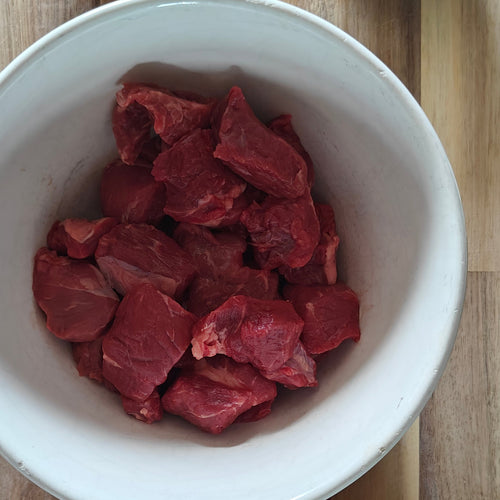 Diced beef in a white bowl on a wooden surface with carrots, an onion, and a small container of spices.