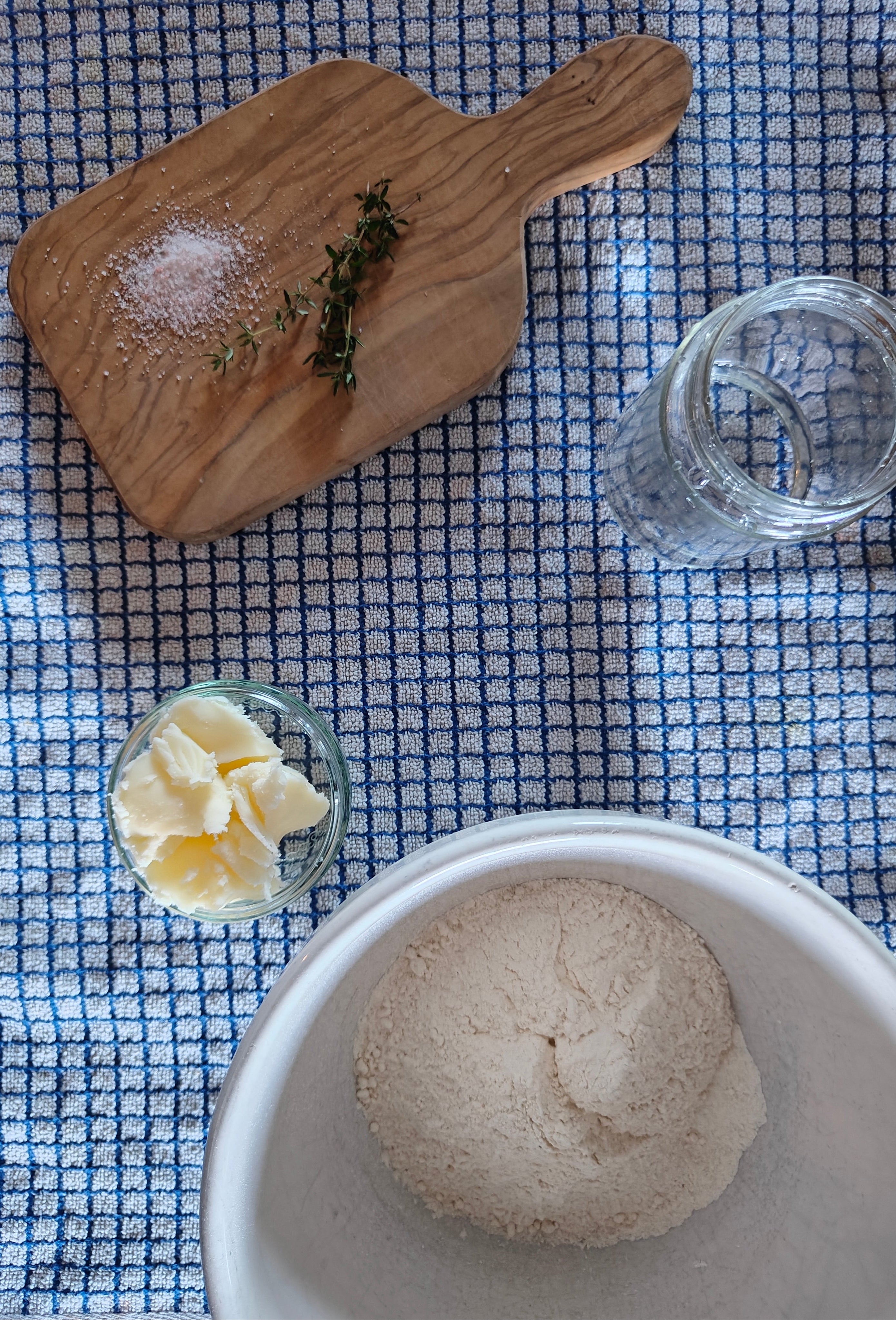 Bowl of flour, glass of water, and wooden cutting board with salt and herbs on a checkered tablecloth.