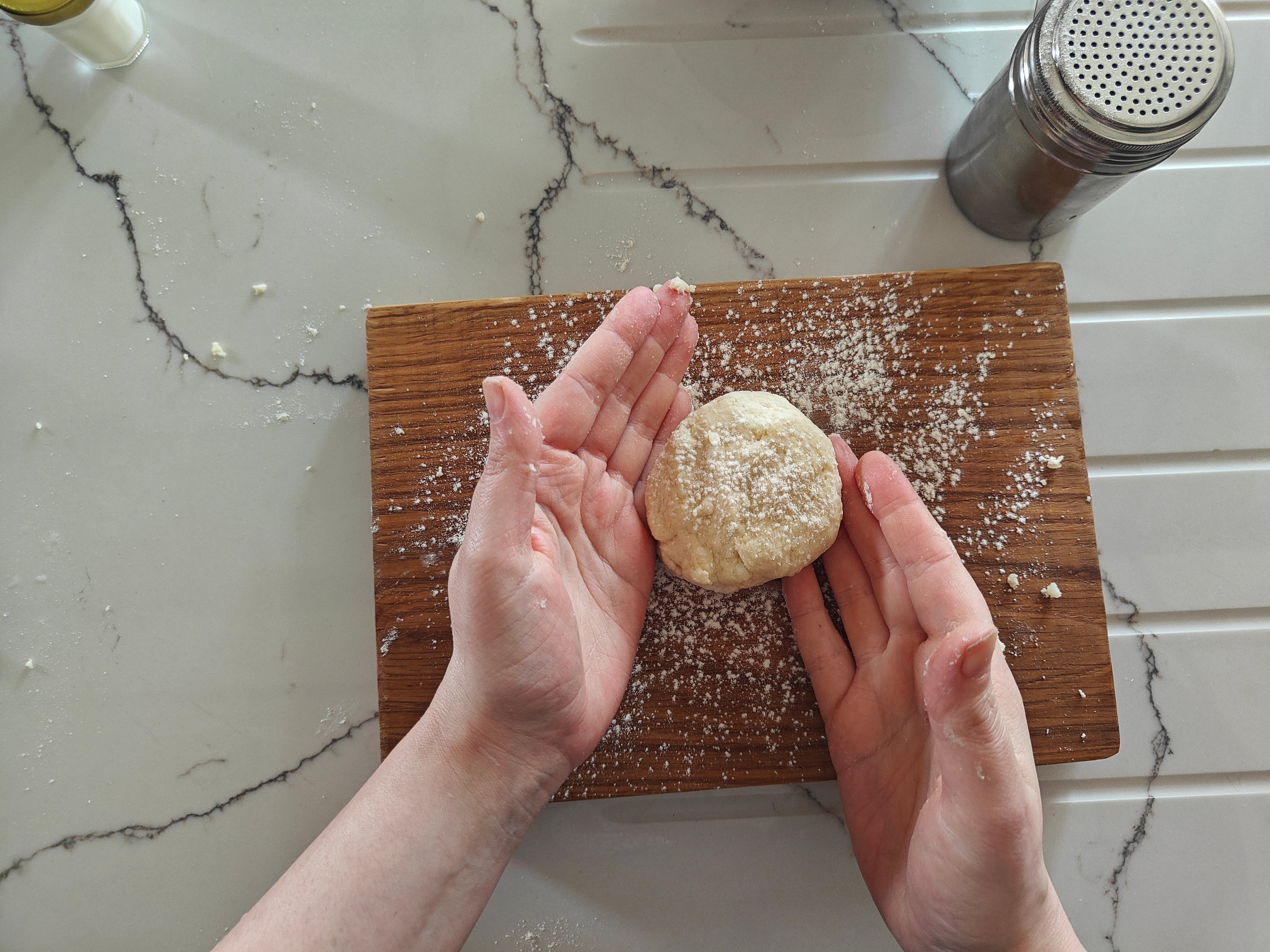 Person holding a ball of dough on a wooden board with flour, against a marble countertop.