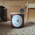 A dark amber glass candle jar with a light wood lid sits on a textured burlap surface in front of a wooden gift box tied with twine. The front label reads 'MELTARA MOISTURE' and 'ROSEMARY & THyme'.