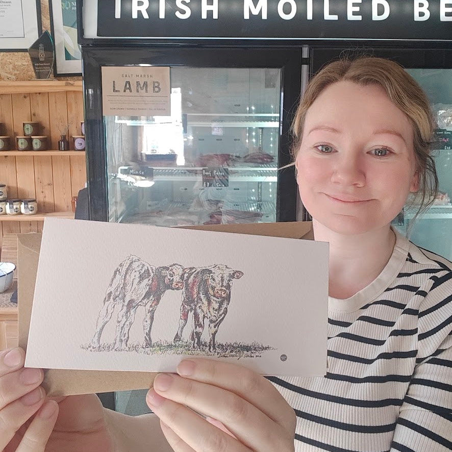 A woman in a black and white striped shirt holds up a printed card featuring a sketch of two cows, with a shop display in the background that reads 'IRISH MOILED BEAR'.