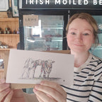 A woman in a black and white striped shirt holds up a printed card featuring a sketch of two cows, with a shop display in the background that reads 'IRISH MOILED BEAR'.