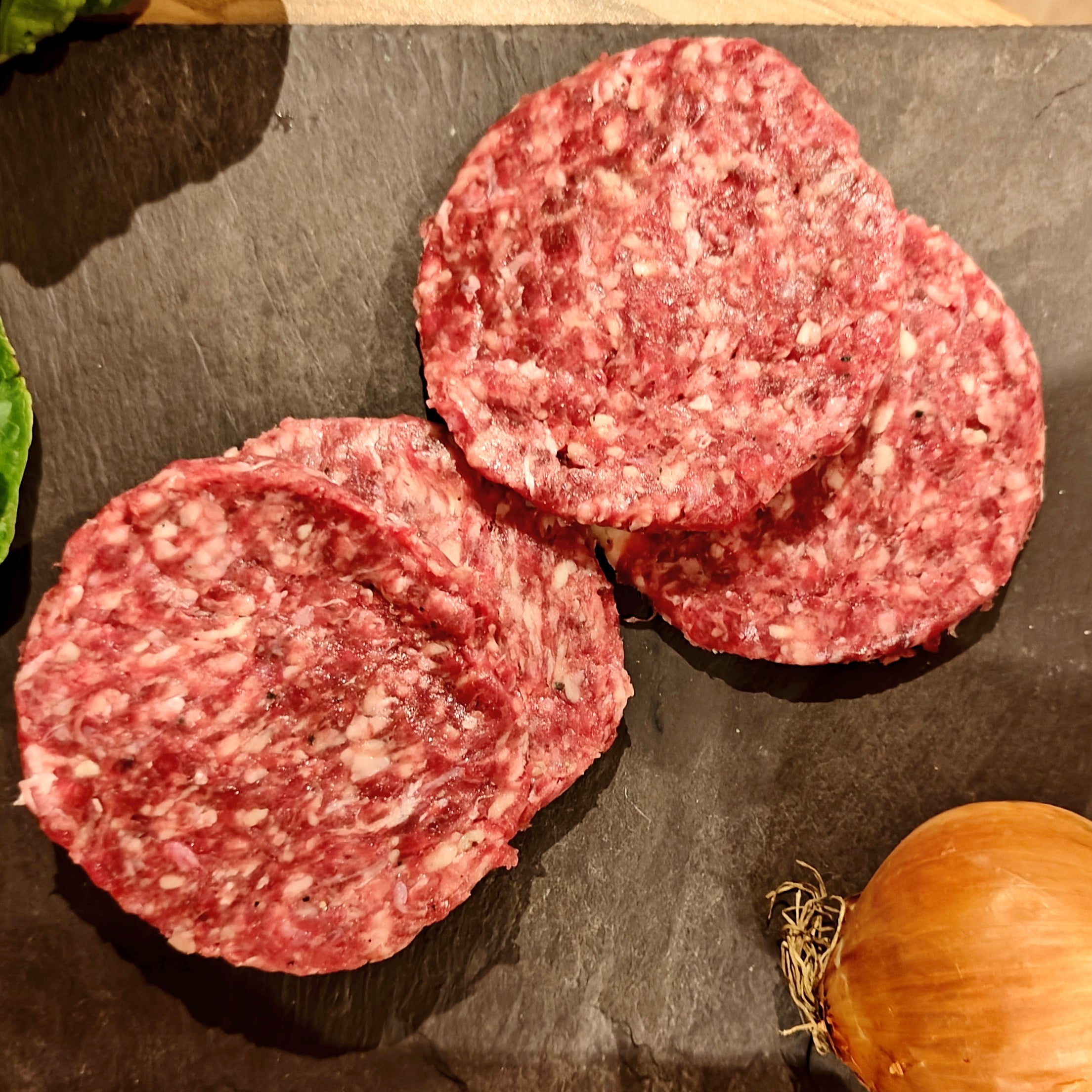 Four raw hamburger patties on a dark surface with lettuce and an onion.